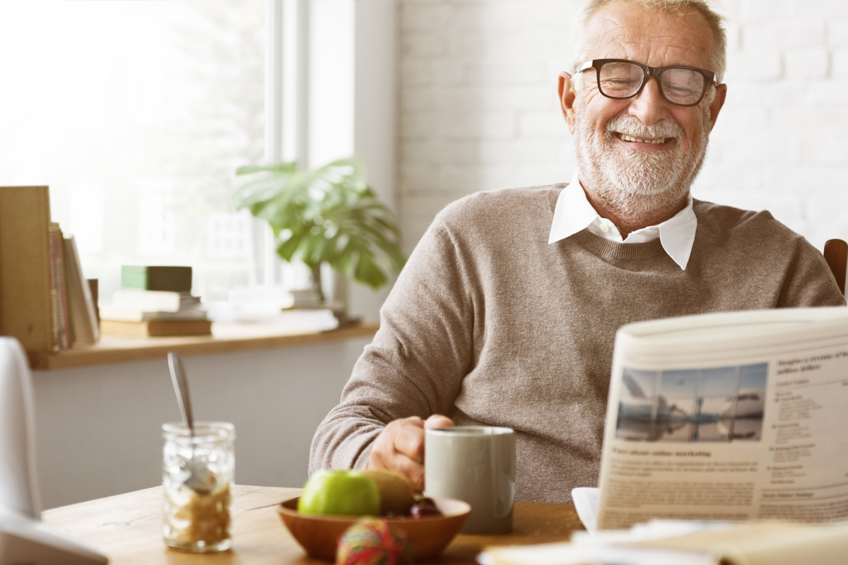 Eldre mann med briller og skjeggstubber sitter ved frokostbordet og leser avisen og holder kaffekoppen