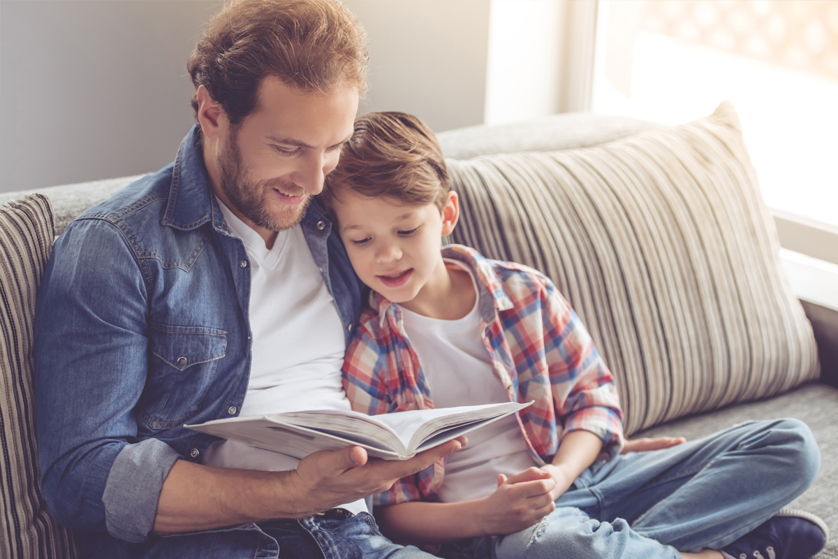 Ung mann med 4 dagers skjegg sitter i sofaen og leser en bok for sønnen