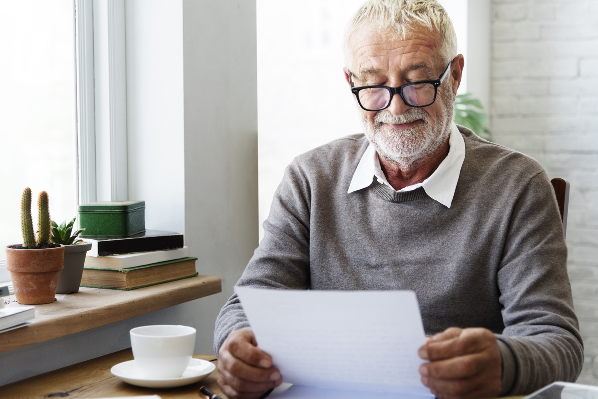 Eldre mann med briller og skjeggstubber leser et brev med kaffekoppen ved siden av